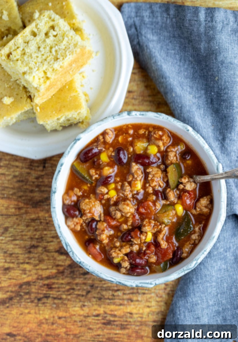 A close-up shot of a bowl of Vegetable Turkey Chili topped with fresh cilantro and shredded cheese, with cornbread in the background.