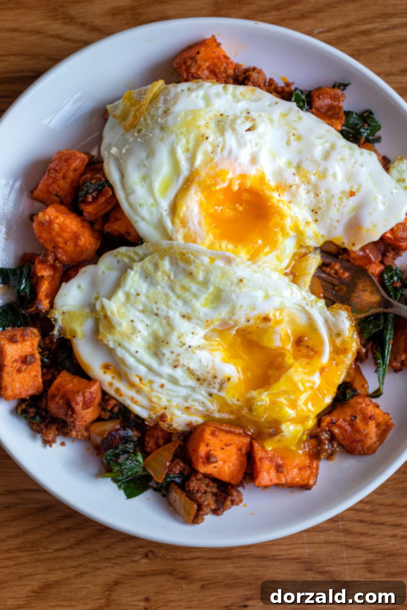 Chorizo Sweet Potato Kale Hash topped with perfectly fried eggs, garnished with fresh herbs, presented on a rustic white plate, emphasizing a healthy and hearty breakfast option.