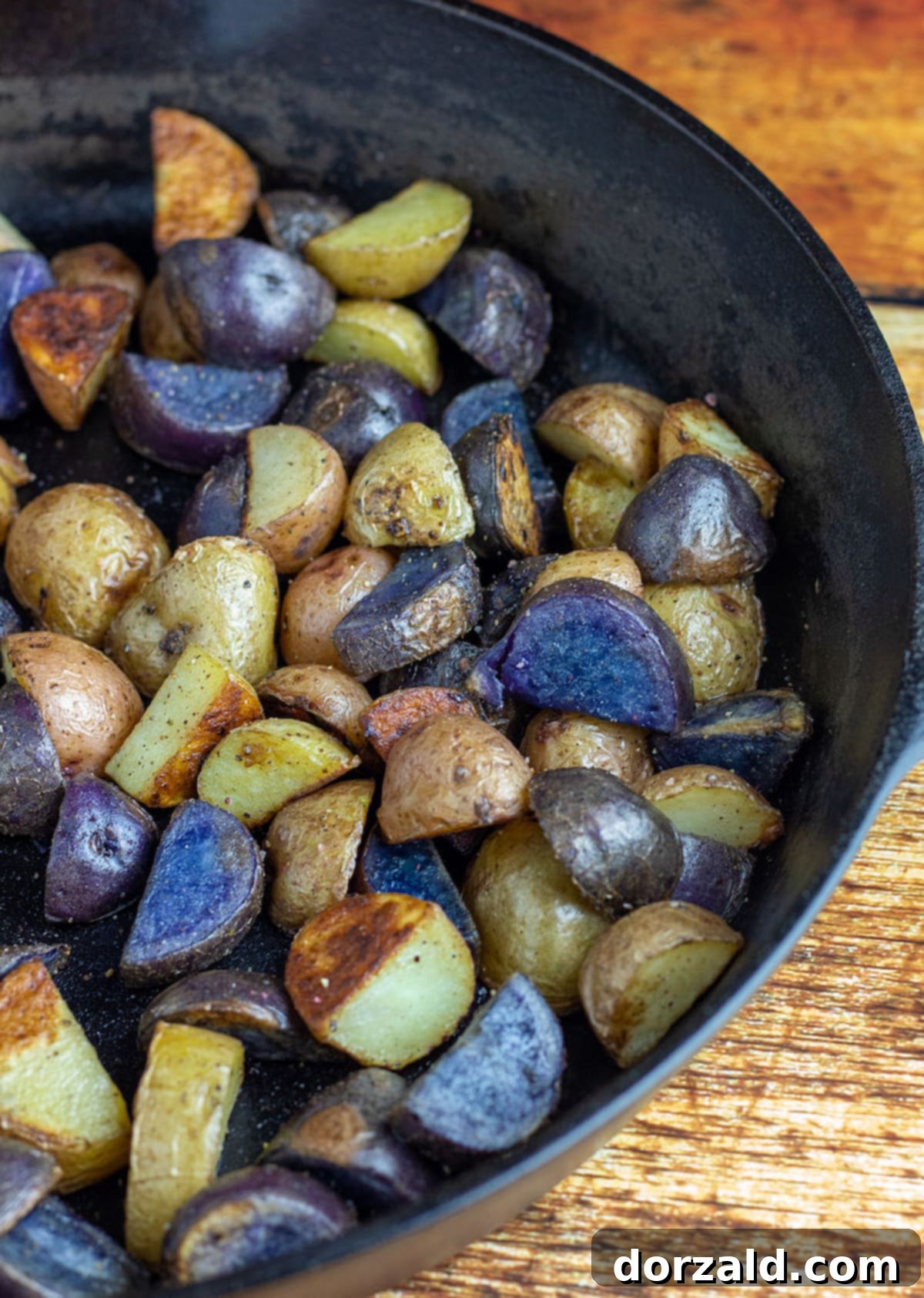 Multi-colored baby potatoes, perfectly roasted to a golden crisp, presented in a large black cast iron skillet on a rustic wooden background.