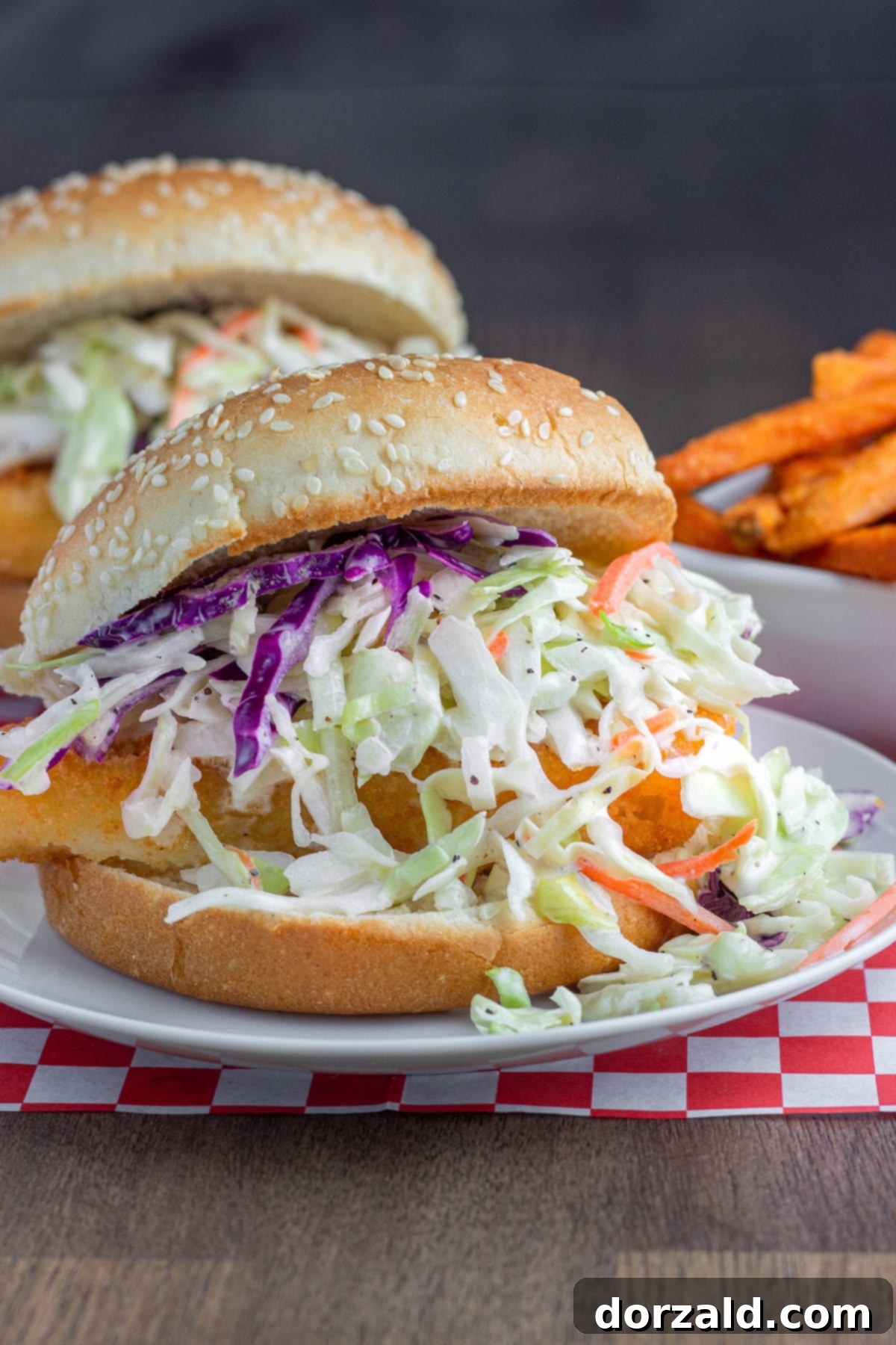 A delicious fish sandwich on a sesame bun, generously topped with vibrant, colorful easy coleslaw. It sits on a red and white checkered napkin, with another similar sandwich and a bowl of sweet potato fries blurred in the background, showcasing the versatility of this easy coleslaw recipe.