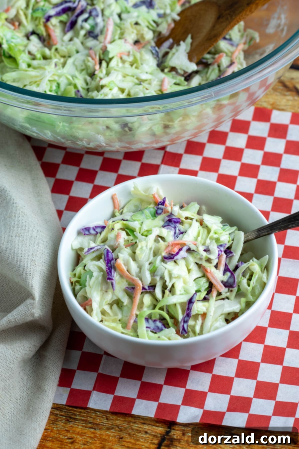 Small bowl of colorful homemade easy coleslaw with a spoon, sitting on red and white checkered paper with a larger serving bowl of coleslaw behind it, ready to be enjoyed.