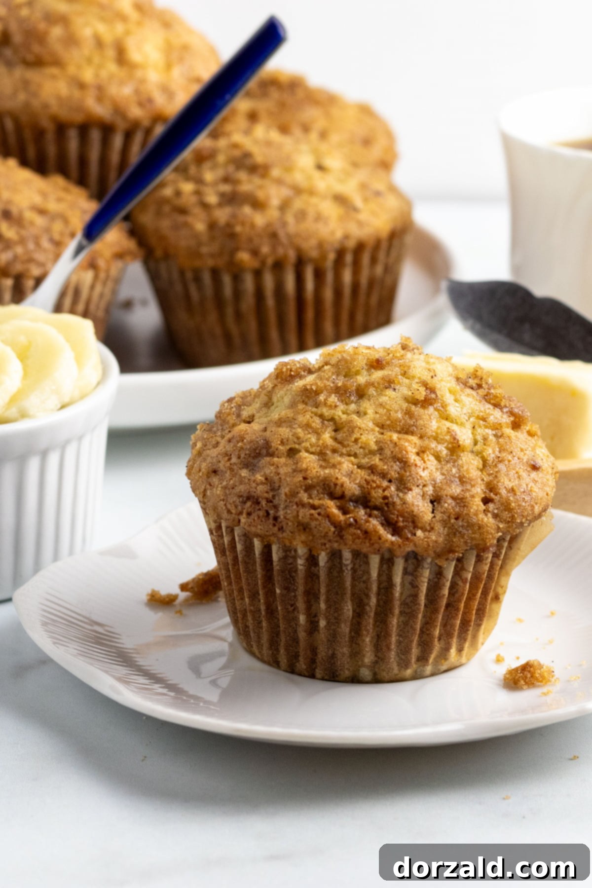 dairy free banana muffin on a small white plate with a plate of muffins, a cup of fruit and a cup of coffee behind it