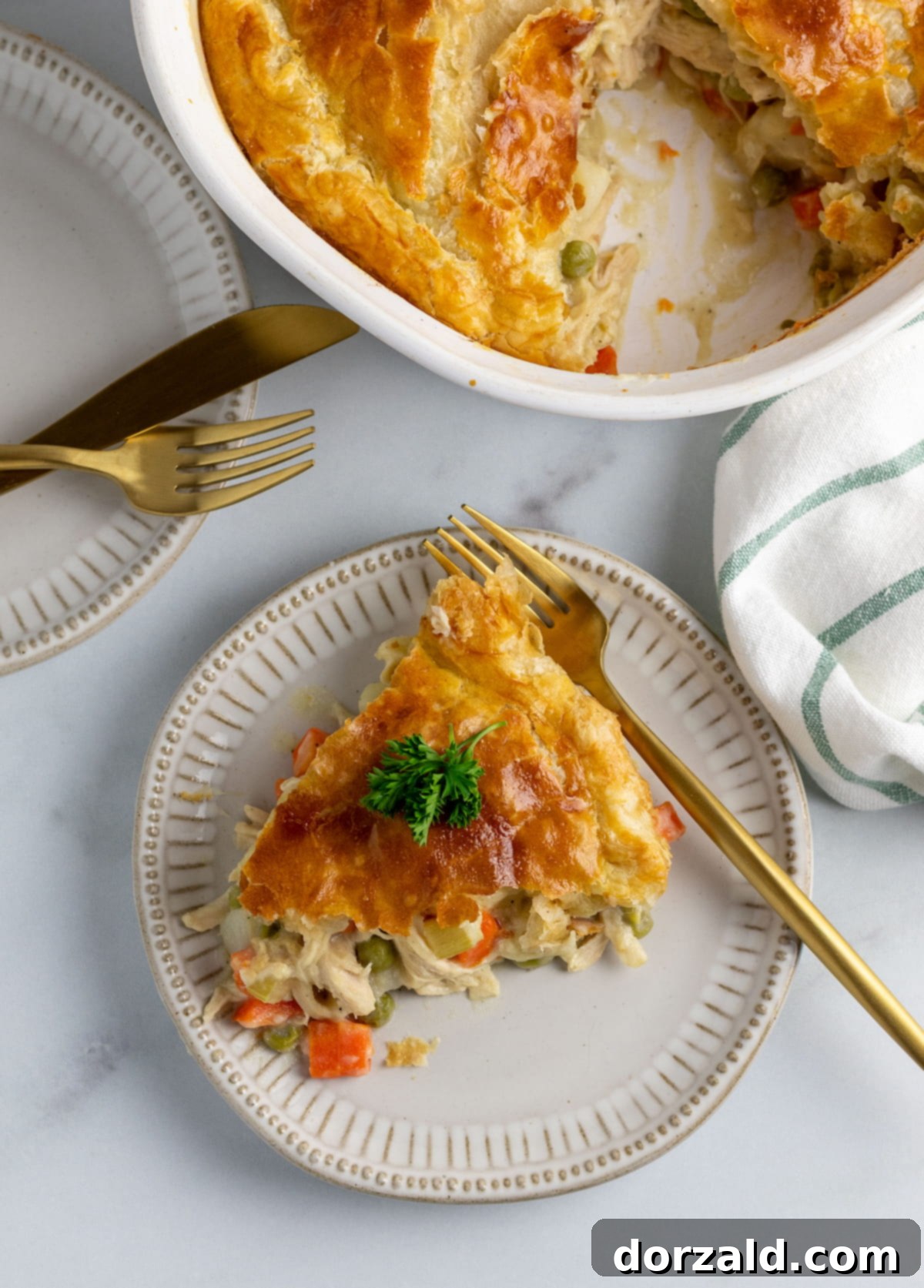 A golden-brown dairy-free chicken pot pie, fresh from the oven, served in a classic white casserole dish on a clean marble countertop. A green and white striped towel and a gold knife and fork are elegantly placed beside it, inviting you to dig in.