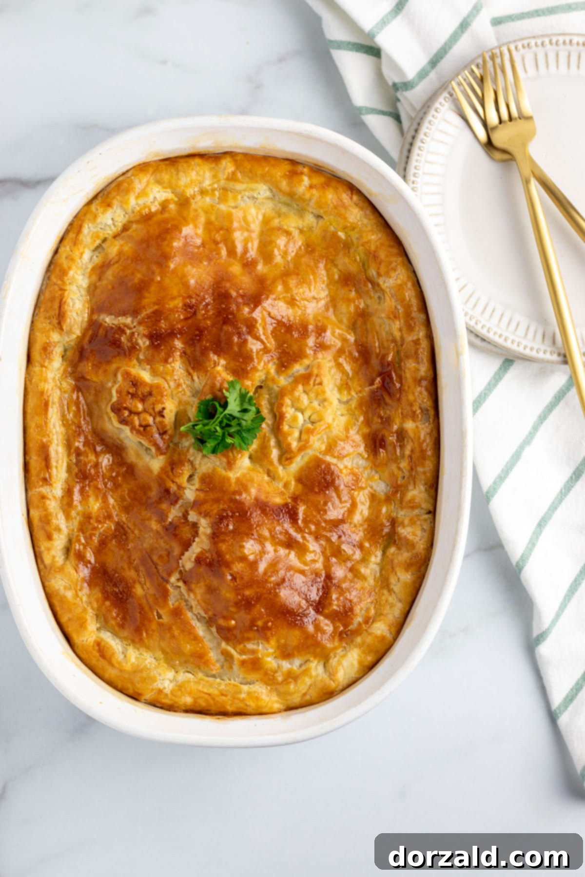 Creamy Dairy-Free Chicken Pot Pie 3 White casserole dish with golden brown dairy-free chicken pot pie, just out of the oven, on a white marble countertop with a green and white striped towel and gold cutlery.