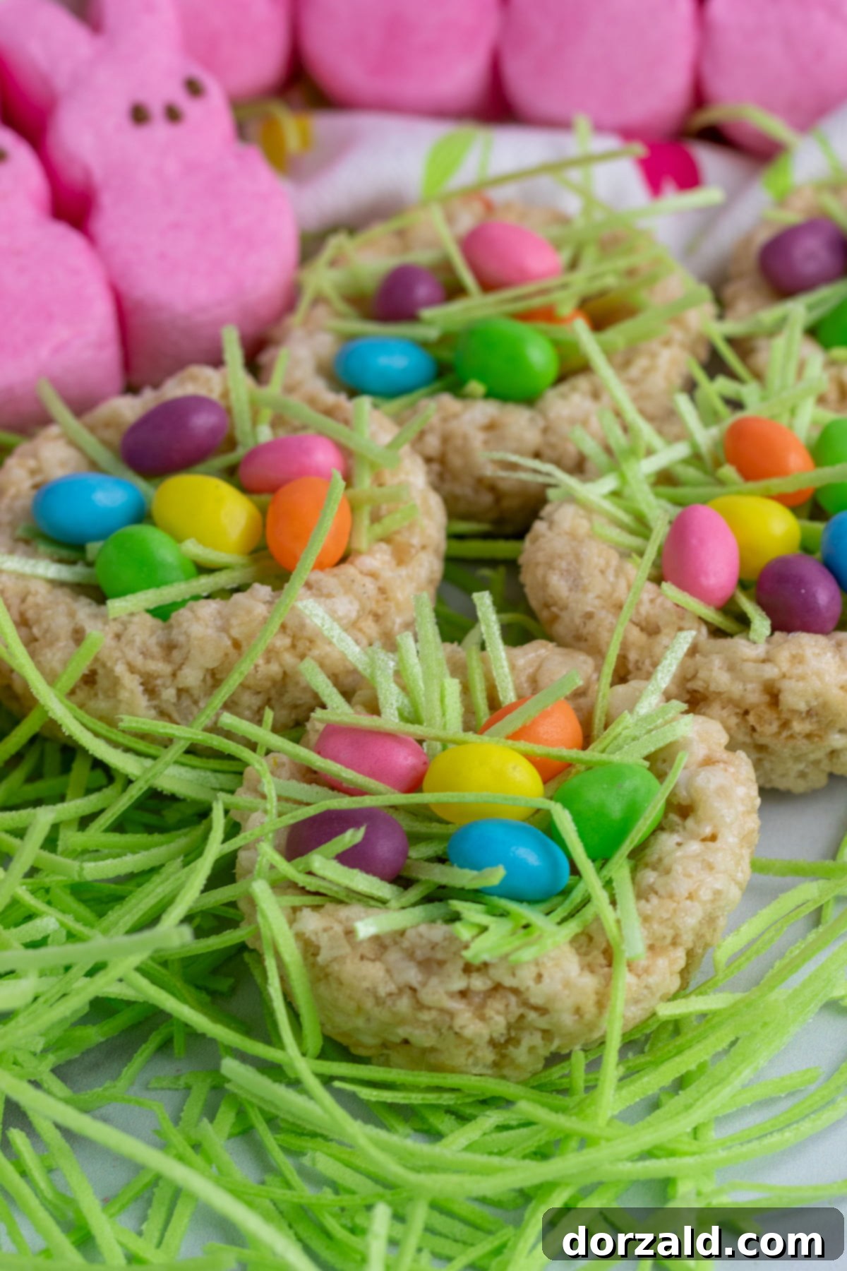 Easy Rice Krispie Easter Nests 2 Close-up of vibrant Rice Krispie Easter Nests filled with colorful jelly bean eggs and edible green grass, with blurred pink marshmallow bunnies in the background.