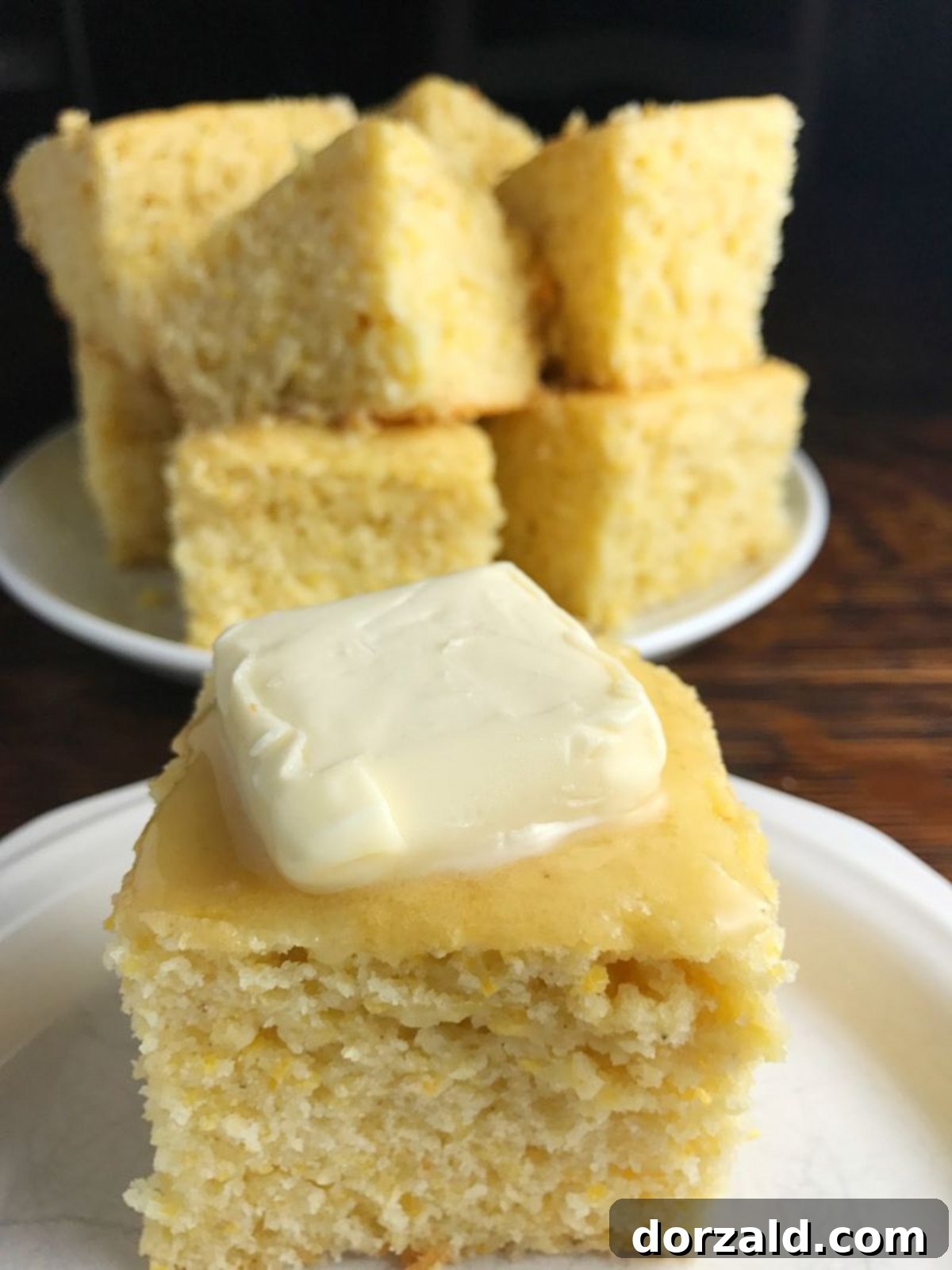 Sweet Dairy-Free Cornbread cut into pieces and stacked on a plate in the background with a single piece on a small white plate in front of it, showcasing its golden-brown crust and soft interior.